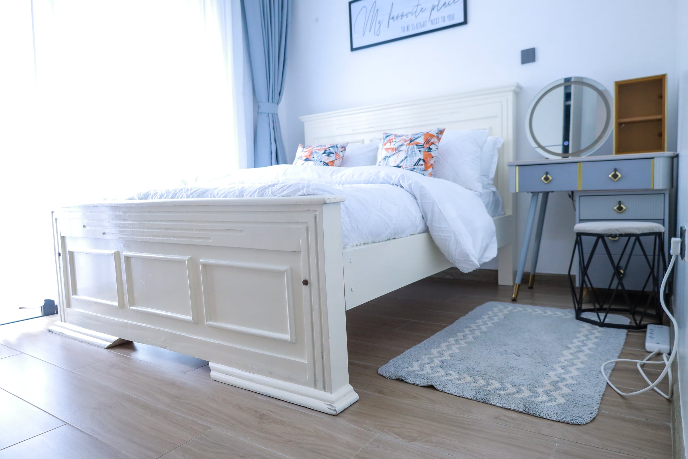 King bed in white cotton beside a blue-grey vanity, round mirror and soft grey rug